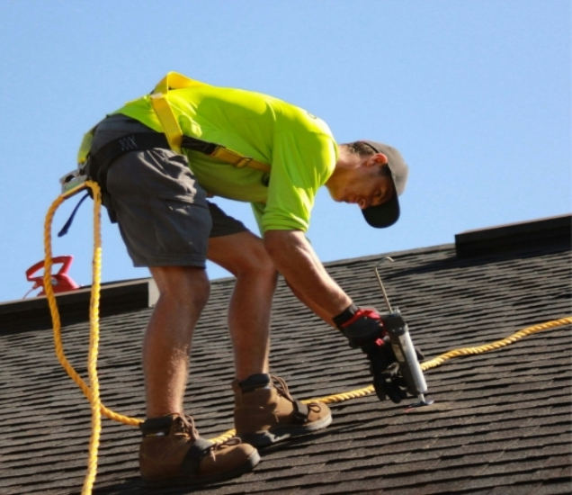 Roofer in a safety harness nailing asphalt shingles on a sloped roof