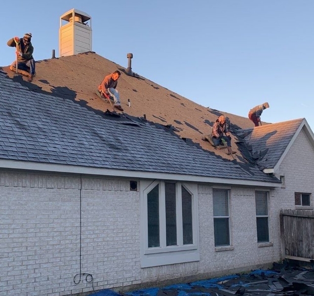 Crew stripping old shingles from the roof of a white brick house