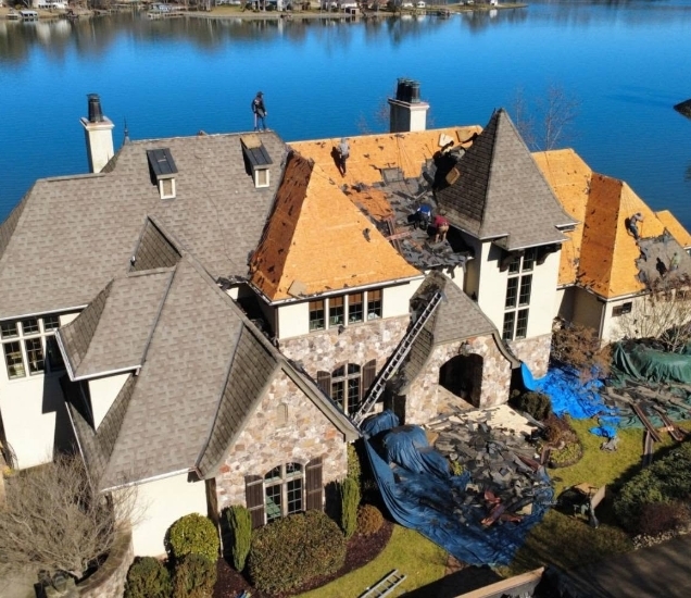 Aerial view of a stone house by a lake with sections of roof stripped to the deck